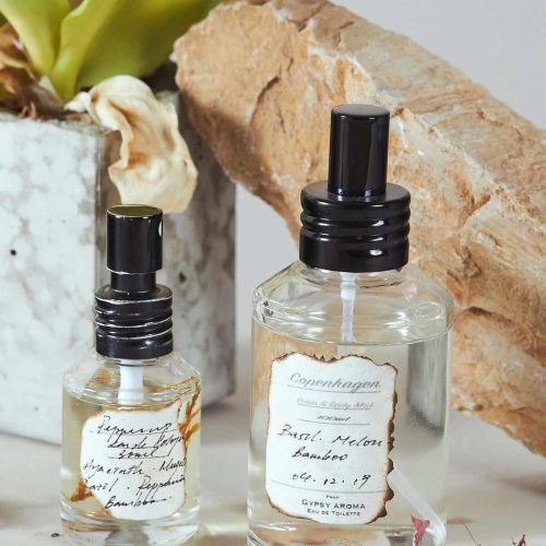 شیشه عطر Ember Gardens Morning Bloom، با برچسب حاوی گل‌ها و پروانه‌ها، بر روی لبه پنجره و در پس‌زمینه گل‌های طبیعی. عطرسازی دستی و طبیعی.