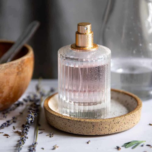 شیشه عطر شیک روی پایه سنگی، با شاخه‌های اسطوخودوس تازه و ظرف چوبی در پس‌زمینه. عطرسازی طبیعی و لوکس.